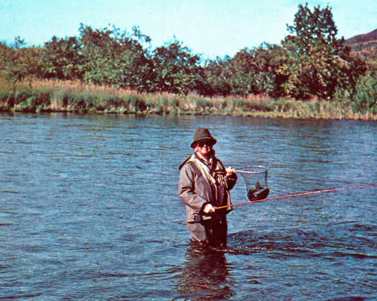 A fly angler standing mid-stream with a steelhead in a landing net.