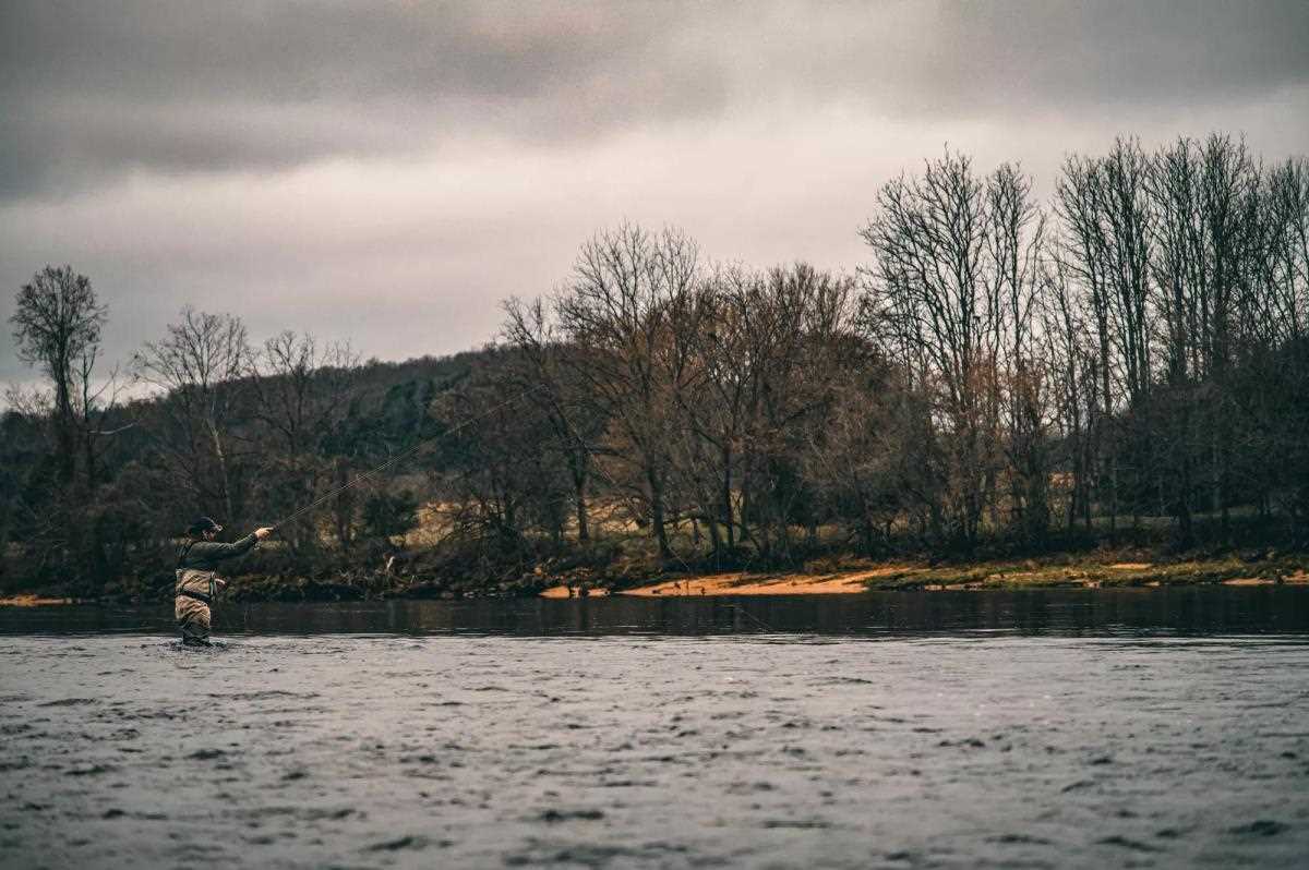 A wading fly angler casting across a riffle with leaf-less trees in the background, on a cloudy day.