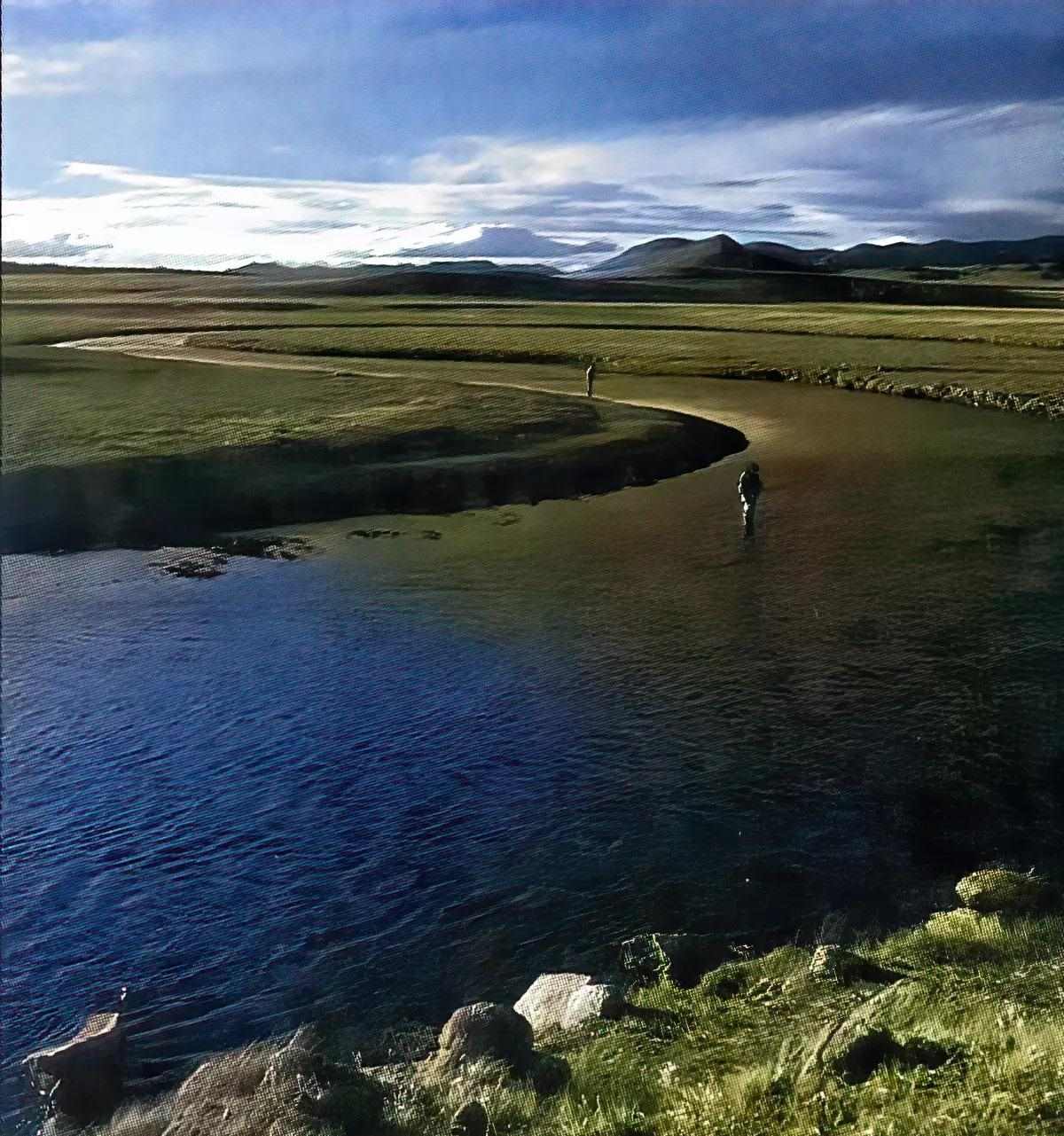 Two fly anglers wade fishing a serpentine bend in a river with mountains in the background. 