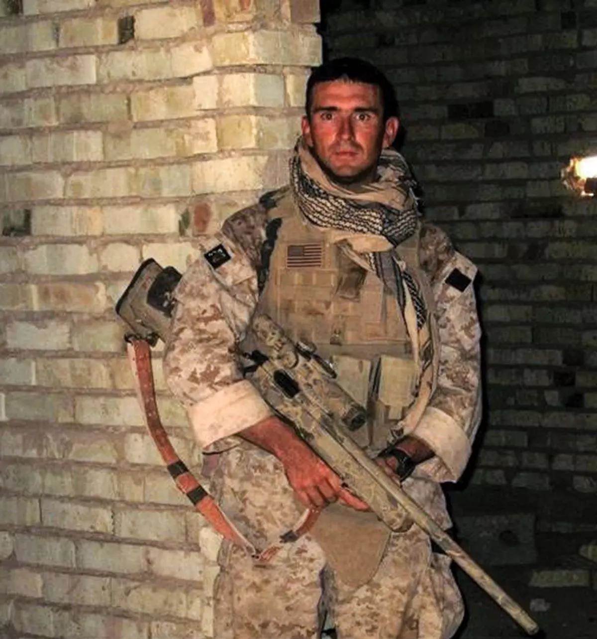A marine in desert camo stands next to a brick wall with his rifle. 