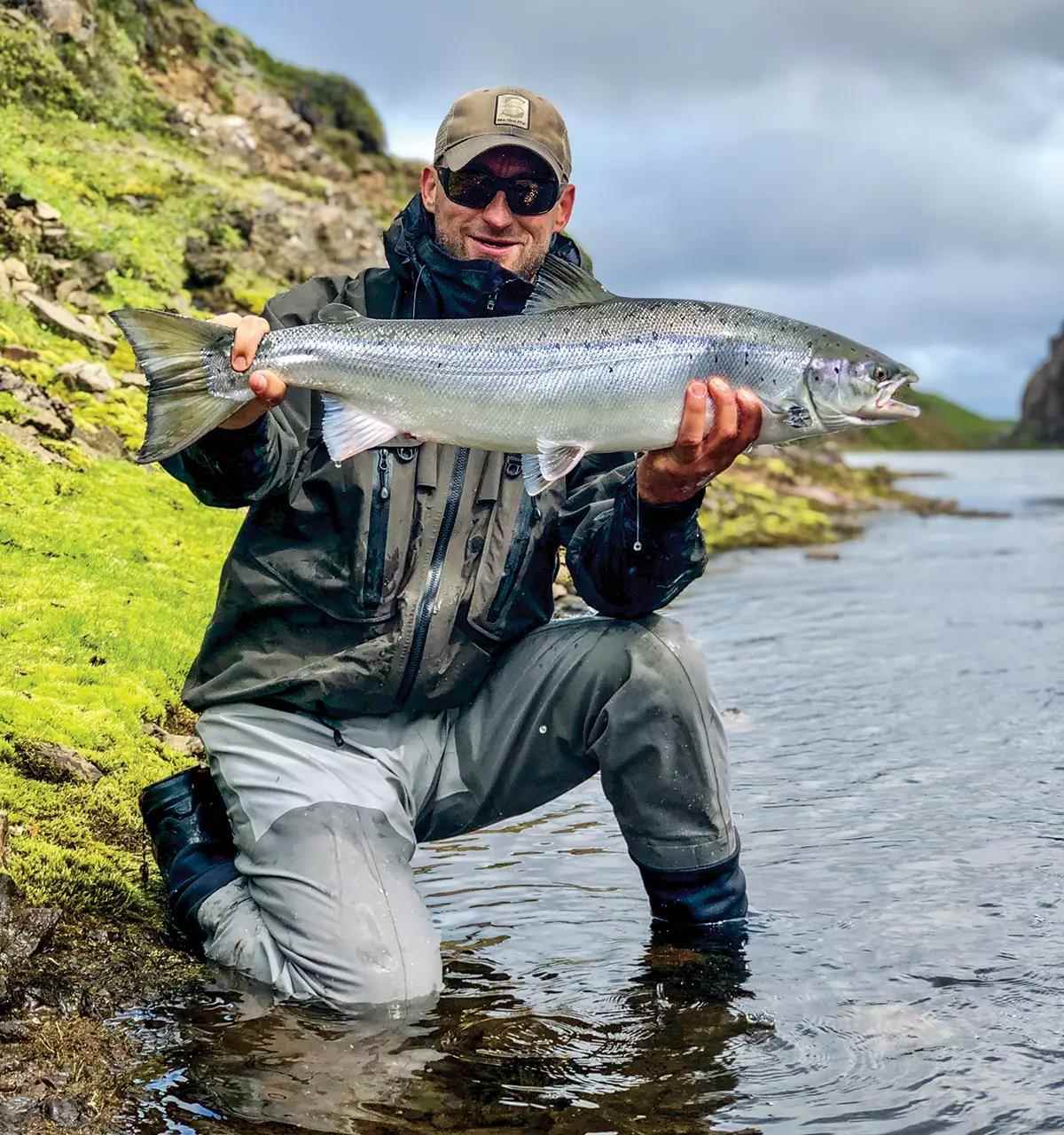 A fly angler kneeling on the bank of a river holding a large Atlantic salmon.