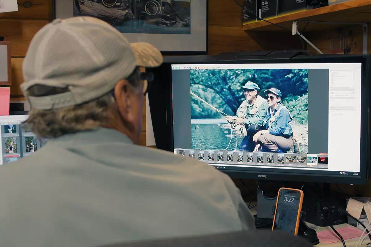 A man in a hat sitting behind a computer screen looking at fishing images.