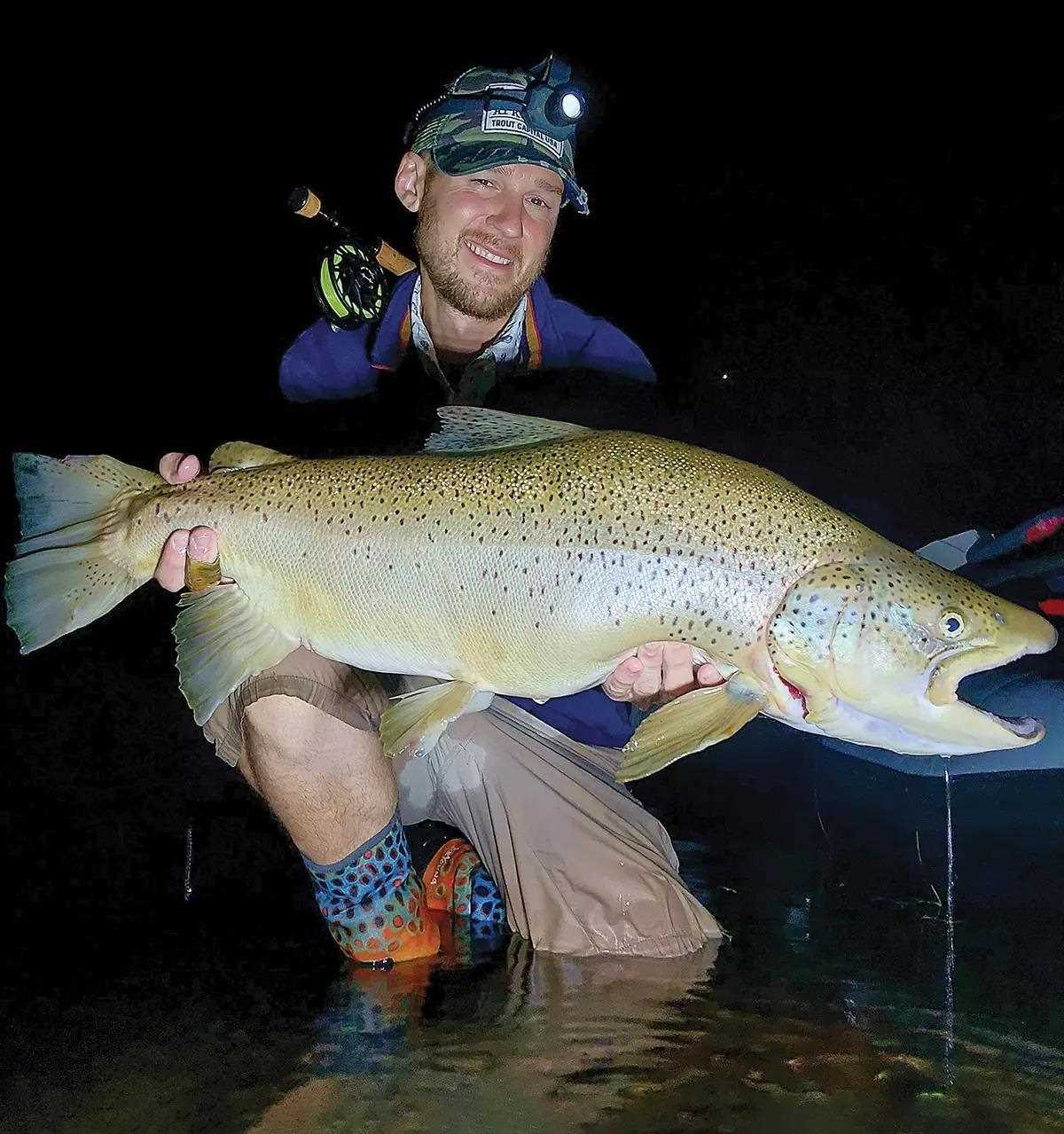 A fly angler holding a huge brown trout for the camera at night. 