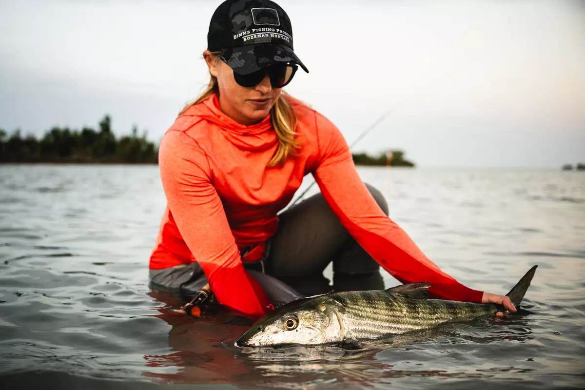 A woman holds a bonefish half in/half out of the water as she squats; a fly rod and reel rest on her lap.