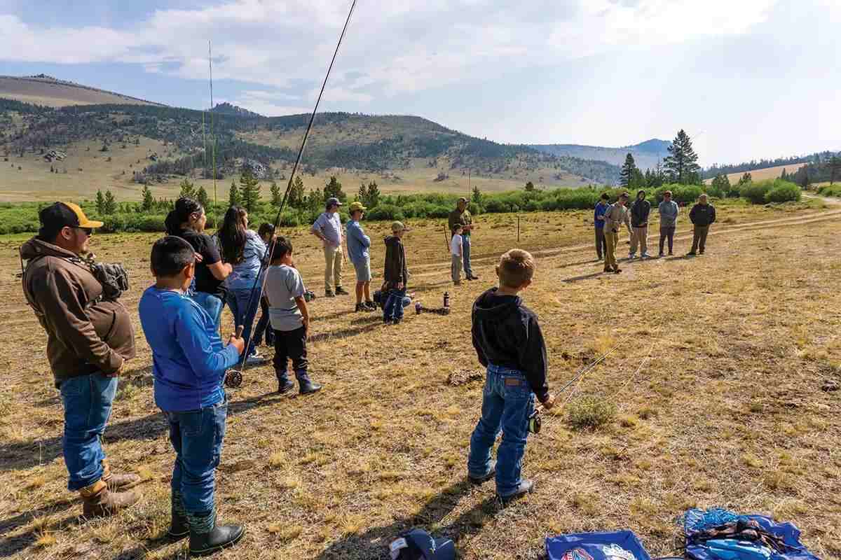Several kids and a few parents watch a fly-casting demonstration in a dry landscape with mountains in the distance.