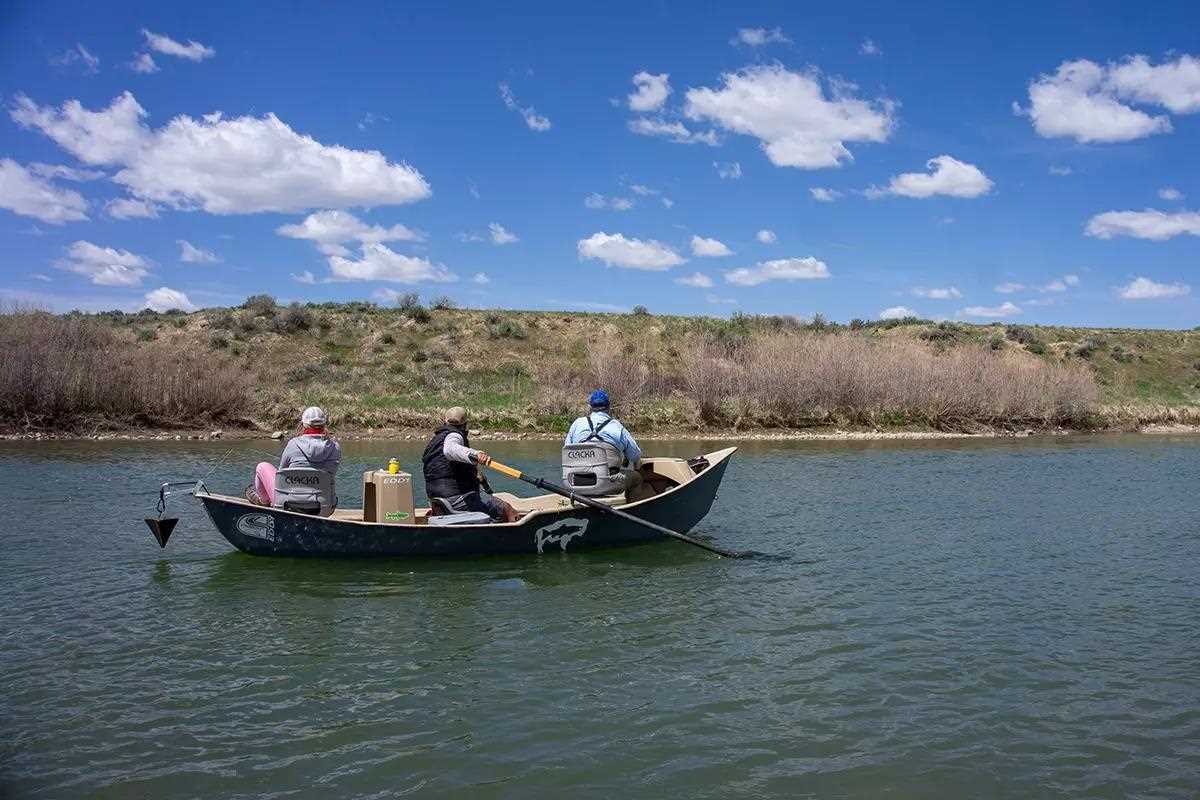 A drift boat with two fly anglers and a fishing guide floating down the river on a sunny day.