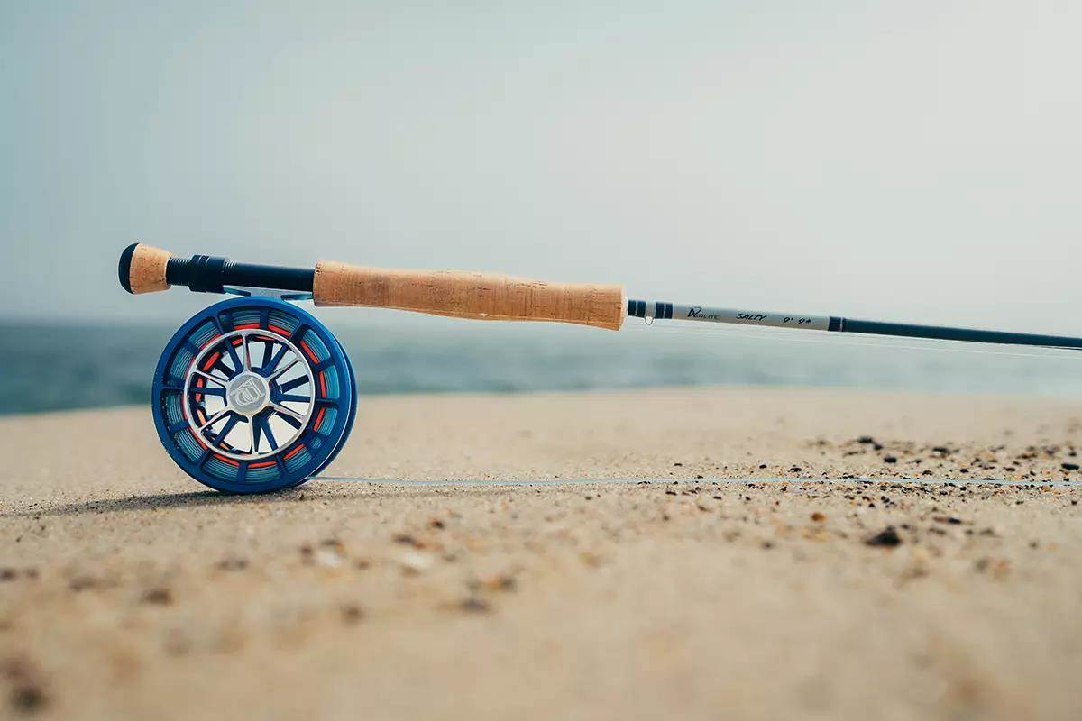 A fly rod and reel sitting on the sand of an ocean beach. 