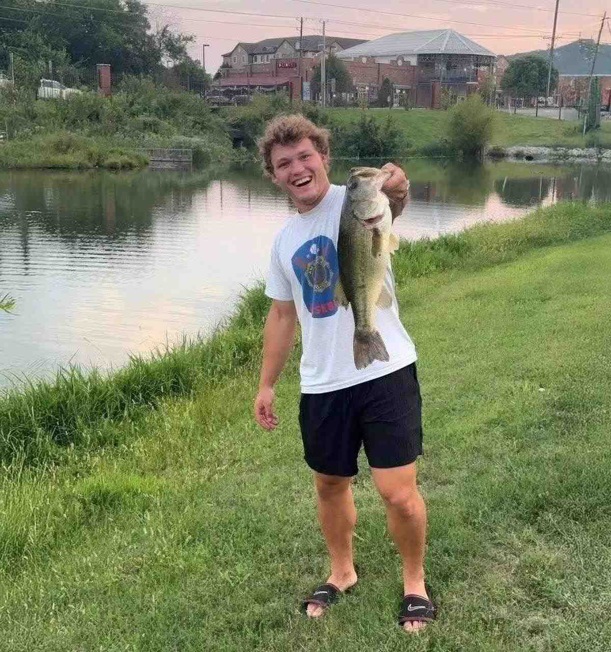 A young man smiles holding a large largemouth bass on the banks of a pond.
