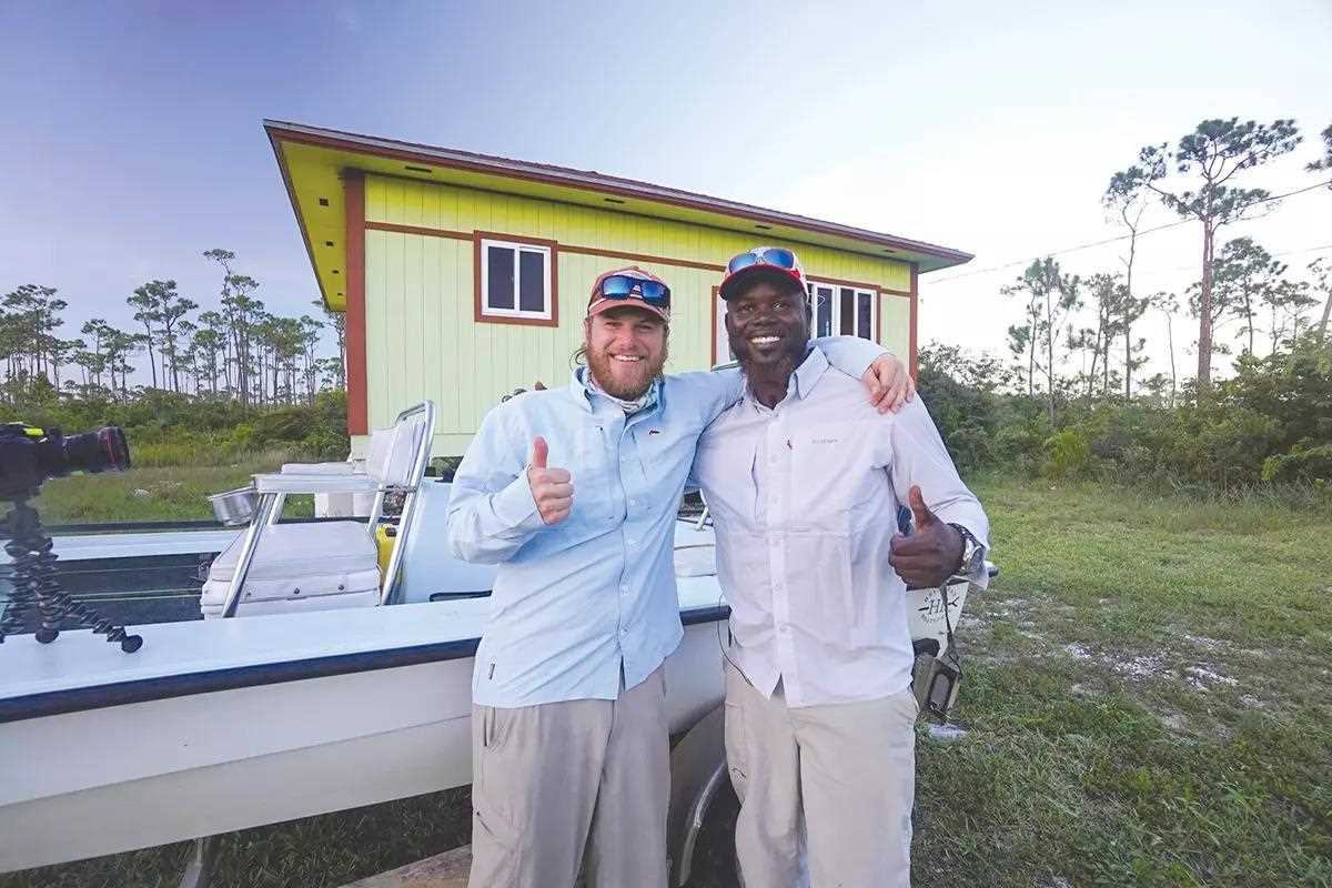 Harrison Buck and Meko Glinton smile for the camera, arm in arm with thumbs up, in front of a flats boat at the Meko Experience.