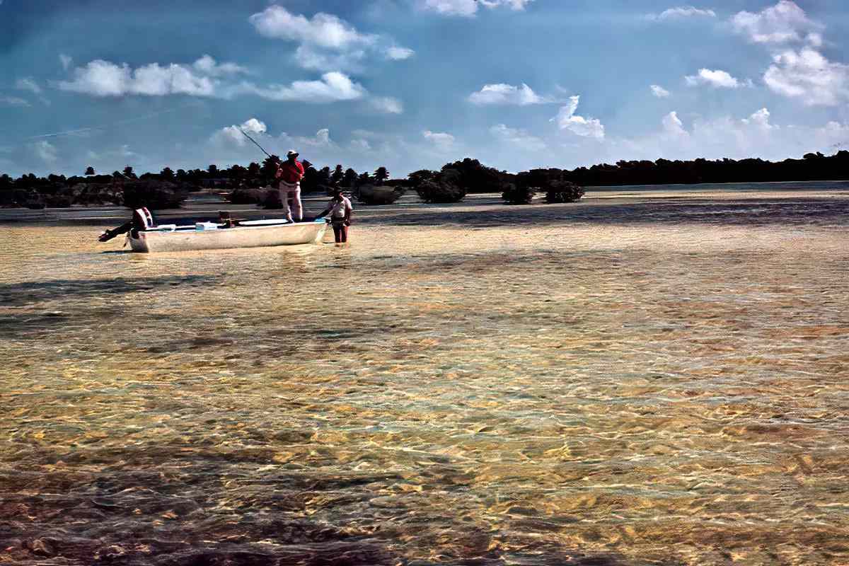 A fishing guide standing in a shallow salt-water flat holding his boat in which a fly angler is casting. 