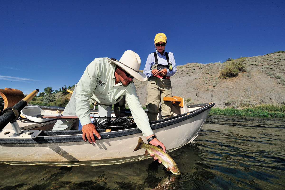 Two fly anglers in drift boat, one leaning over gunwale releasing a trout, the other watching behind; Bighorn River float fishing